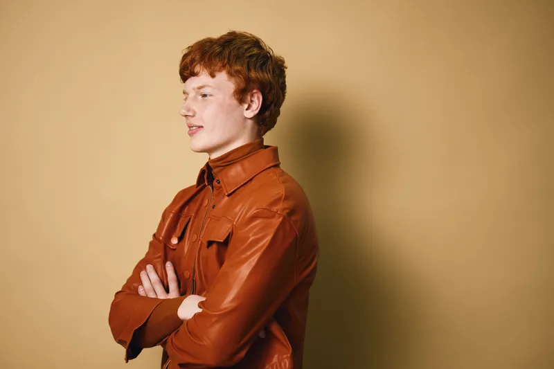 Young man with red hair in brown leather jacket standing against yellow background with arms crossed and looking sideways with confident expression and casual style.