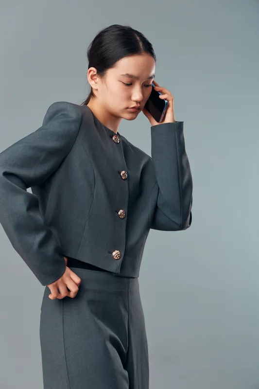 Woman in gray suit talking on phone in thoughtful pose, standing against plain background. Businesswoman with professional attire communicating on mobile device in studio setting.