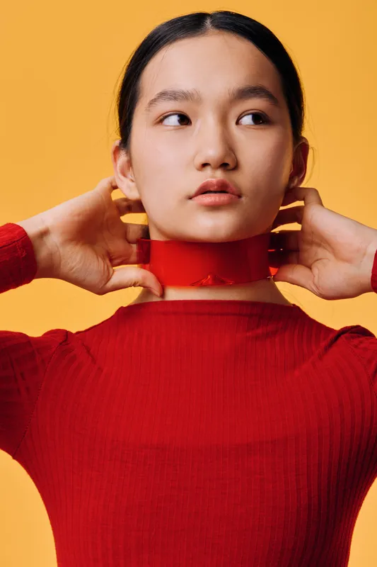 Young woman with dark hair wearing red sweater and red transparent neck accessory standing against yellow background. Portrait of female model with smooth skin and serious expression.