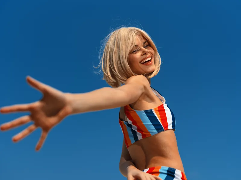 happy young woman in colorful striped swimsuit reaching out with hand, smiling brightly against clear blue sky, summer outdoor portrait with joyful expression