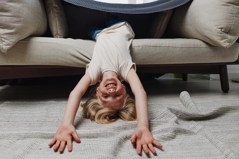 Happy child with blonde hair lying upside down under a couch stretching arms forward on a patterned carpet. Playful kid smiling indoors with casual clothes in a cozy living room environment.