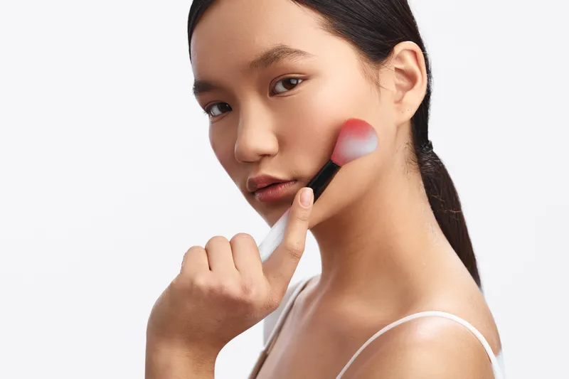 Asian woman applies makeup with a soft brush in a bright studio, capturing a calm, focused moment that highlights personal care, beauty routine, and contemporary self expression.