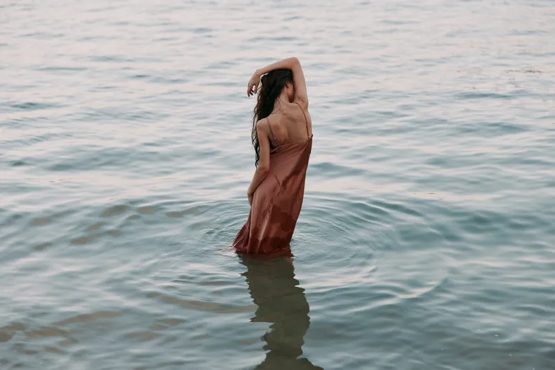 A lone figure stands waist-deep in calm ocean water, wearing a flowing dress as fabric ripples with the tide, creating a serene, poetic shoreline moment.