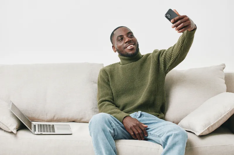 Happy young Black man taking a selfie on a sofa, wearing a green turtleneck sweater, with a laptop beside him, conveying a sense of joy and relaxation