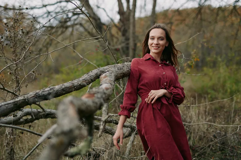 A candid portrait of a woman in a burgundy dress standing near a weathered branch in a wild field, conveying authenticity and quiet confidence with genuine presence.