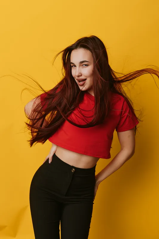Young woman with long hair wearing red crop top and black pants posing with playful expression on yellow background. Fashion, beauty, casual style and confident attitude in studio portrait.