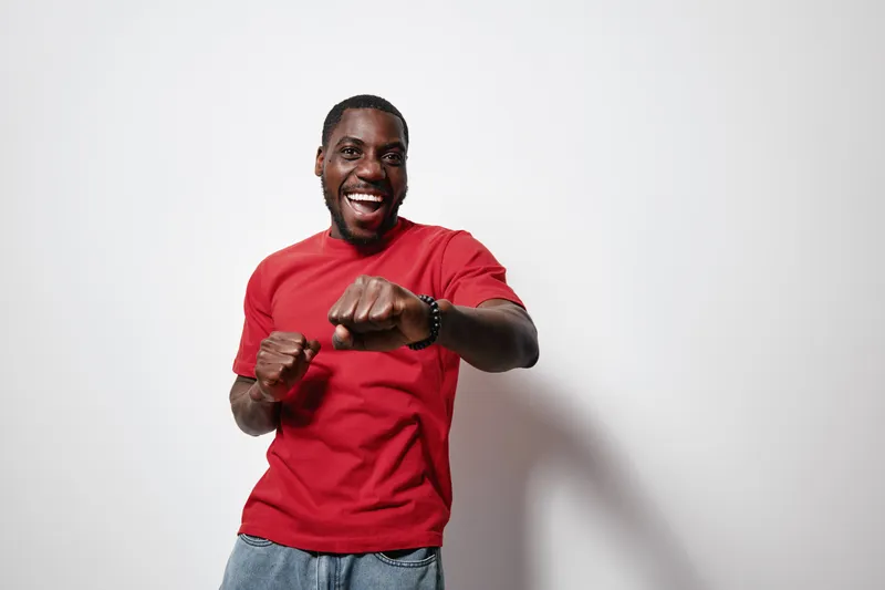 African American man with dark skin wearing red t shirt and jeans smiling and posing with fists up on white background for positive energy and confidence.