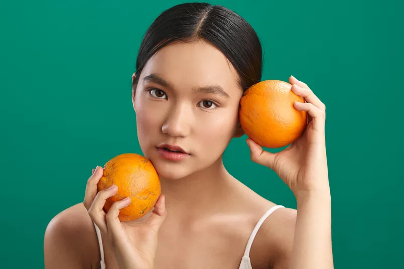 asian woman holds bright oranges close to her face in a studio portrait, creating a vibrant, fresh mood with soft lighting, bold color, and a modern aesthetic.