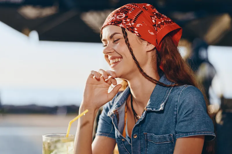 smiling woman in red bandana enjoying sunny day outdoors with refreshing drink by the waterfront, casual denim outfit, happy and relaxed mood