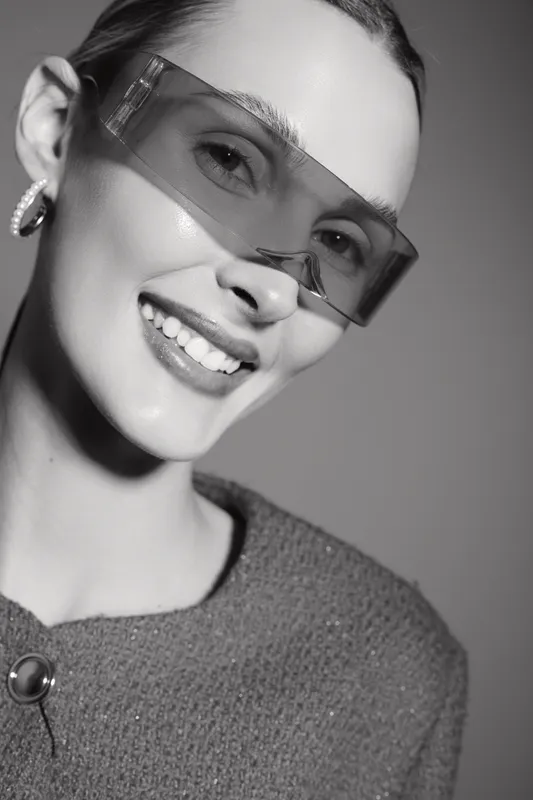 Smiling white woman with fair skin wearing oversized transparent sunglasses and hoop earrings in soft studio light.