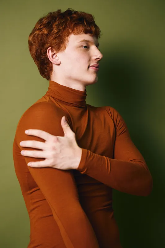 Young man with red hair wearing brown turtleneck stands in profile with arm crossed touching opposite shoulder on green background. Casual fashion portrait with confident expression.