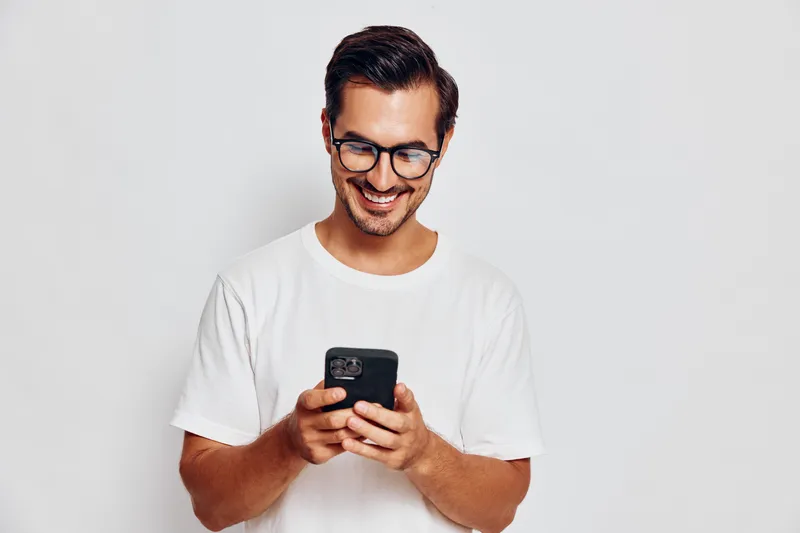 Smiling young man wearing glasses holds smartphone, looking at screen with cheerful expression isolated on plain light background for lifestyle concept.
