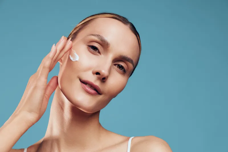 Woman beauty skincare cream portrait studio blue wellness face apply, calm expression, hand touching cheek, soft light, smooth skin, minimal makeup, background