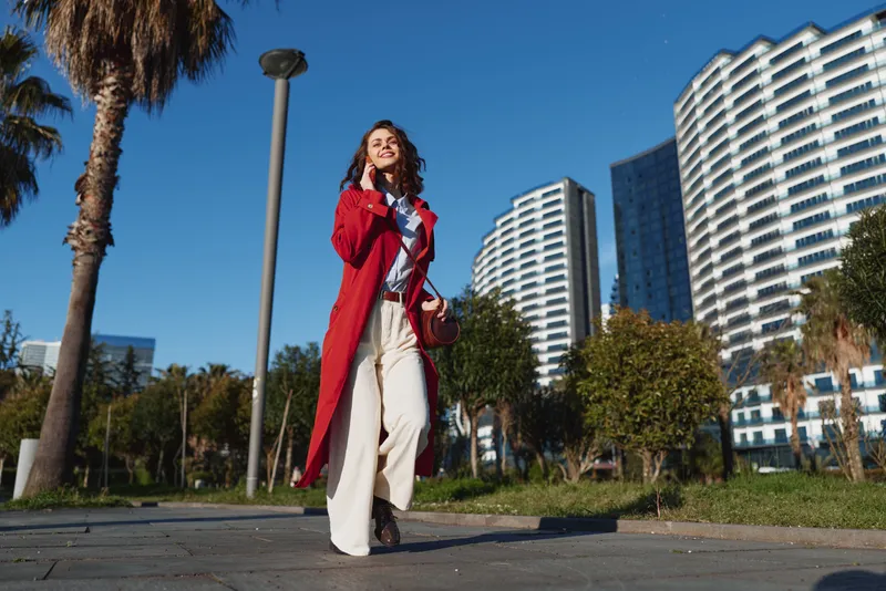A young woman in a red coat walks confidently along a sunny city avenue, with modern high rises behind her, creating a stylish, energetic urban lifestyle moment