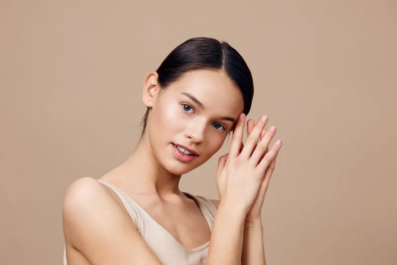 Young woman with a serene expression, wearing a beige sleeveless top, hands gently clasped on a soft beige background, conveying calmness and beauty