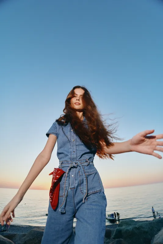 young woman in denim jumpsuit posing outdoors near sea with sunset sky and red bandana, casual style and relaxed mood in warm evening light