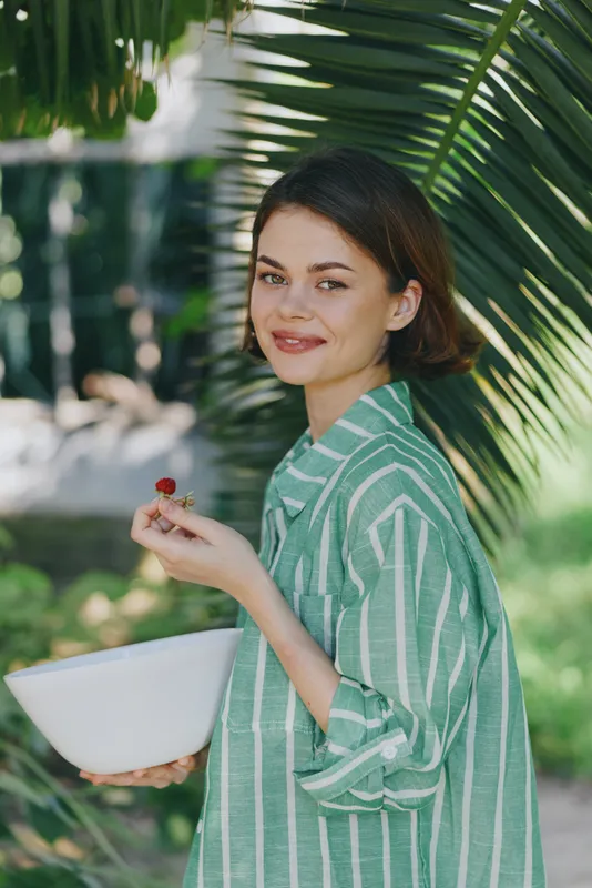 Portrait of a smiling woman in a striped green shirt holding a white bowl and a small berry, standing beside tropical palm leaves in bright outdoor light.