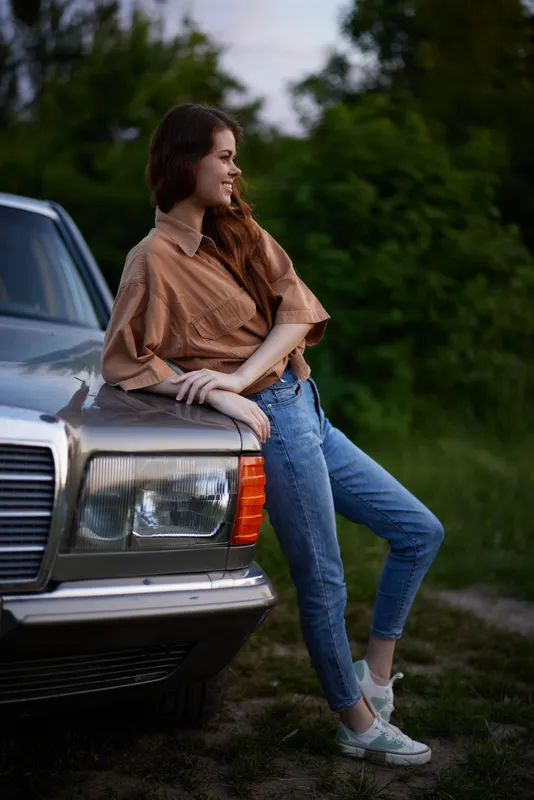Young woman wearing a stylish outfit leaning on a vintage car in a lush green setting, conveying a relaxed summer vibe with a nostalgic touch.