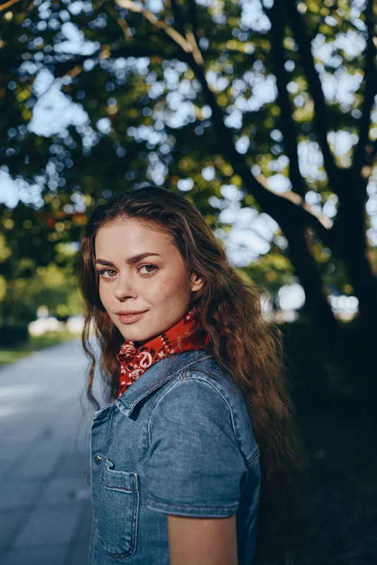 young woman in denim shirt outdoors with curly hair and red bandana smiling softly under green trees in warm natural light