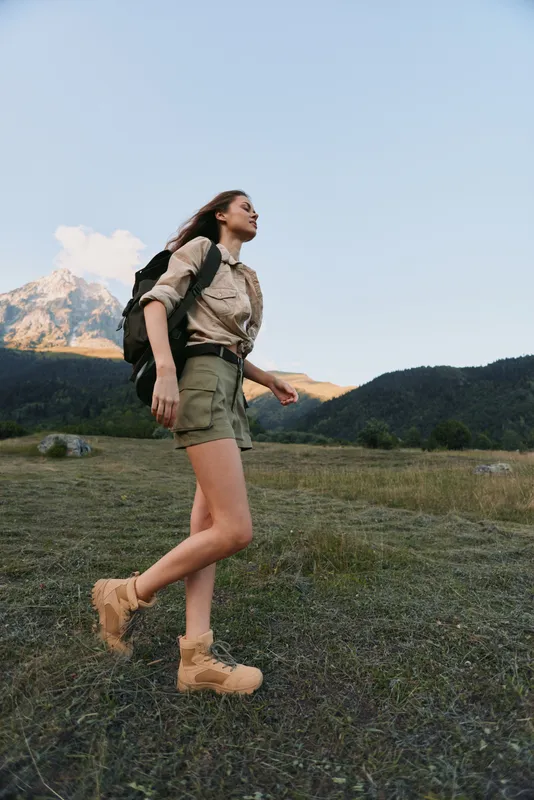 Woman hiking in a natural landscape with a backpack and boots, wearing casual outdoor attire. Wide field with distant mountains, fresh air, and an adventurous mood.