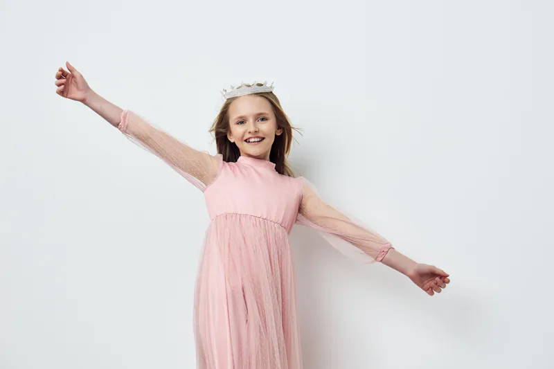 Happy girl in a pink dress with sheer sleeves, wearing a crown, celebrating joy on a light background, perfect for children s fashion concepts