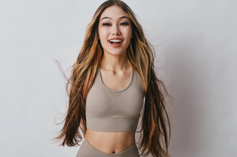Young woman with long hair wearing sportswear smiling and posing in studio. Female athlete with light makeup showing happiness and confidence in activewear outfit for fitness training.