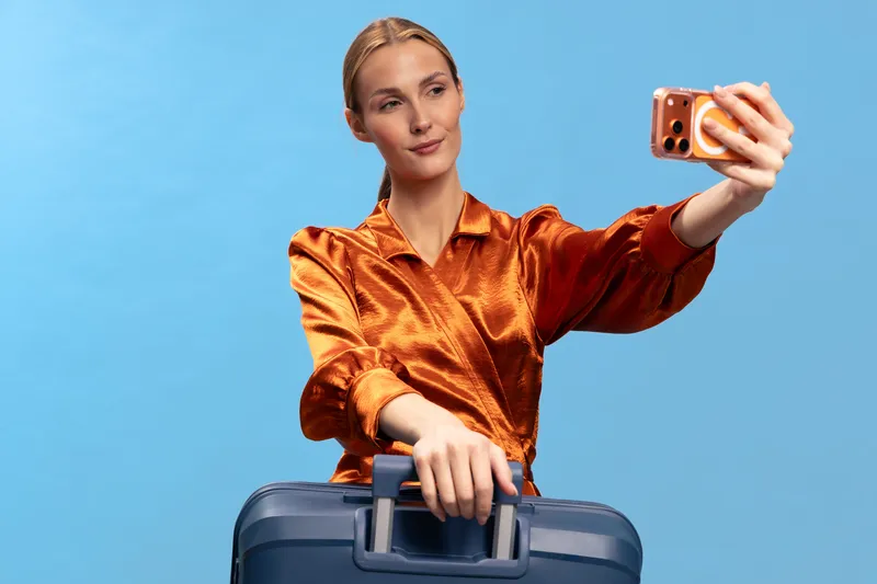 Young woman in orange blouse takes selfie with smartphone while holding suitcase handle. Blue background highlights travel concept, tourism, vacation, and modern lifestyle with casual outfit.
