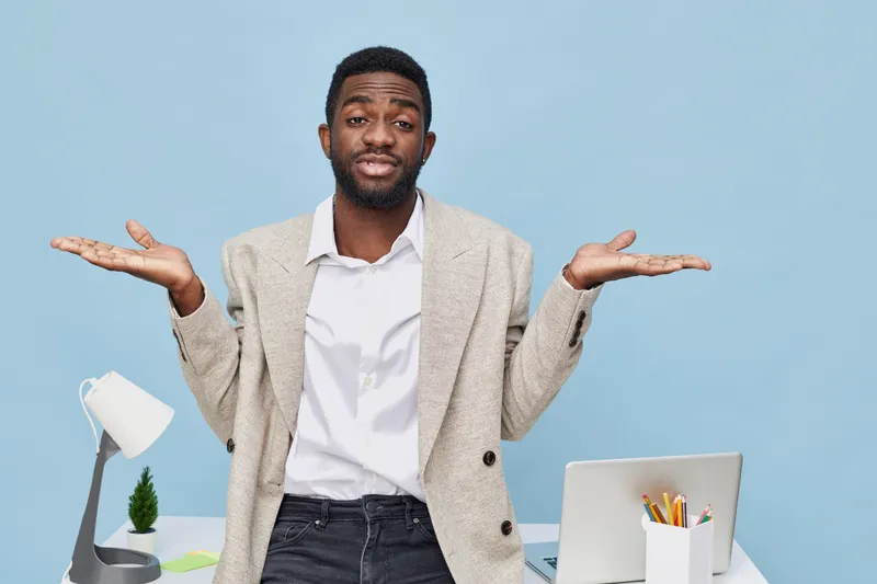 Confused man with raised shoulders and open palms wearing business casual outfit, standing behind desk with laptop, lamp, and office supplies isolated on blue background. People concept, workplace dil