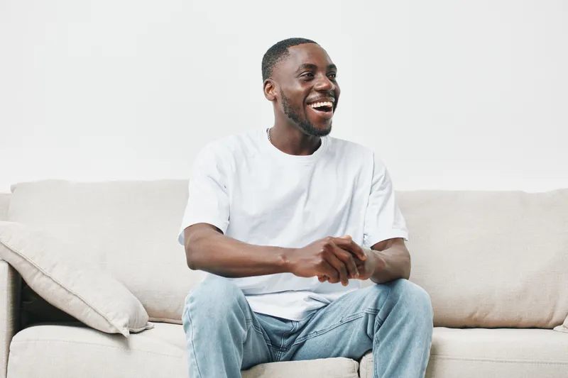 Happy young African American man sitting on a beige sofa, wearing a white t shirt and jeans, expressing joy and relaxation in a modern, bright interior