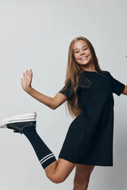 Girl jumping and smiling in black dress and sneakers with long hair, energetic happy child posing on white background in casual style with sport socks and joyful expression.