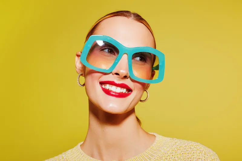 Smiling woman with bright red lipstick wearing large blue sunglasses and yellow sweater on yellow background. Concept of fashion, summer style, beauty, and cheerful mood.