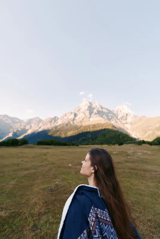 Woman in profile standing in a grassy meadow with expansive mountain landscape, nature scene for travel and outdoors adventure, long hair, patterned sweater and serene panoramic view.