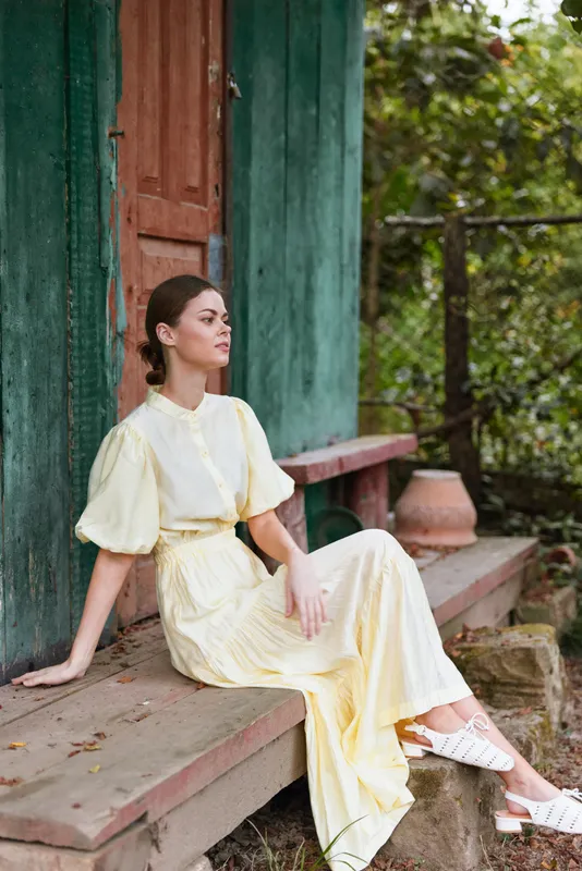 Elegant woman in a yellow dress sits on a rustic porch, serene and composed, with garden and warm outdoor light creating a tranquil moment