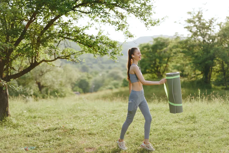 Young woman walking in a green nature setting while carrying a yoga mat, dressed in athletic wear, representing healthy lifestyle and outdoor fitness