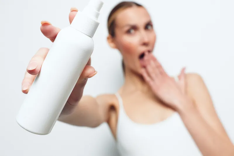 Blurry woman in white tank top holds white spray bottle close to camera, surprised expression, isolated on white background, skincare concept, product focus, minimal style.