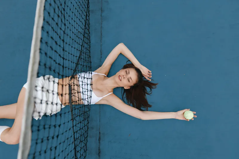 Young woman in sportswear relaxing on a tennis court with a tennis ball after playing, showcasing athleticism and a serene expression on a bright blue background