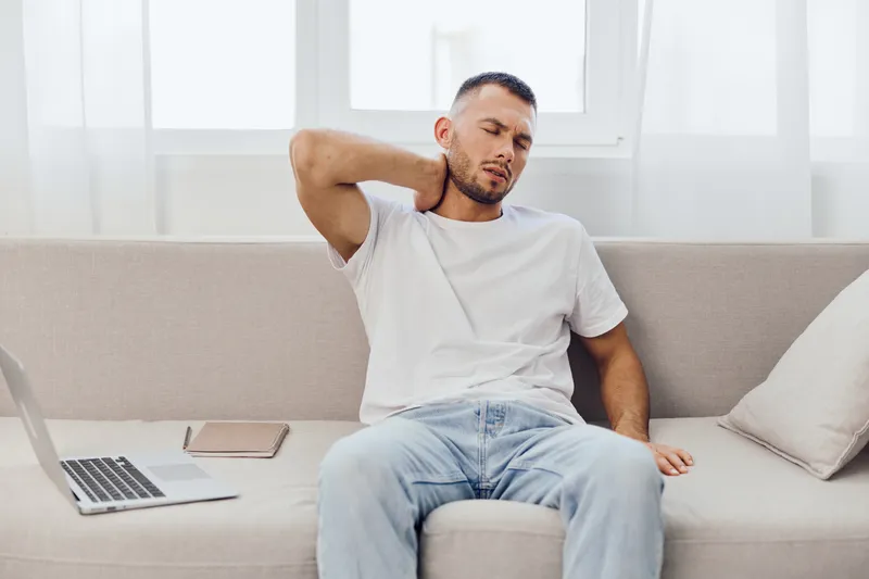 A stressed man sitting on a couch, experiencing work-life blur, showcasing anxiety and fatigue from digital overload. The scene reflects emotional minimalism and vulnerability.