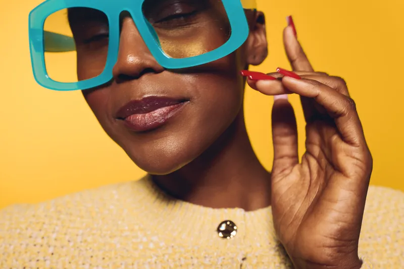 Close-up of a smiling black African woman wearing large blue sunglasses and a yellow sweater. She has dark skin and red nails, posing against a bright yellow background. Concept of fashion and style.