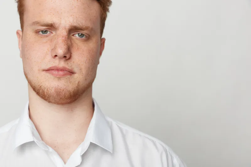Close-up portrait of serious young man with red hair and beard wearing white shirt against plain light background. Concept of confidence, youth, and natural appearance.