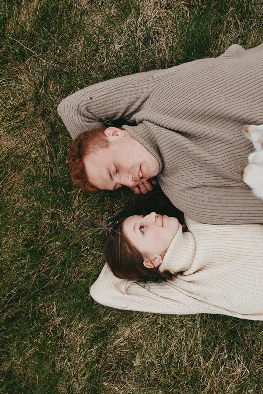 Couple lying on grass wearing cozy sweaters, sharing a relaxed outdoor moment, warm light and calm mood, lifestyle vibe, candid connection, natural textures, intimate pause in a breezy day