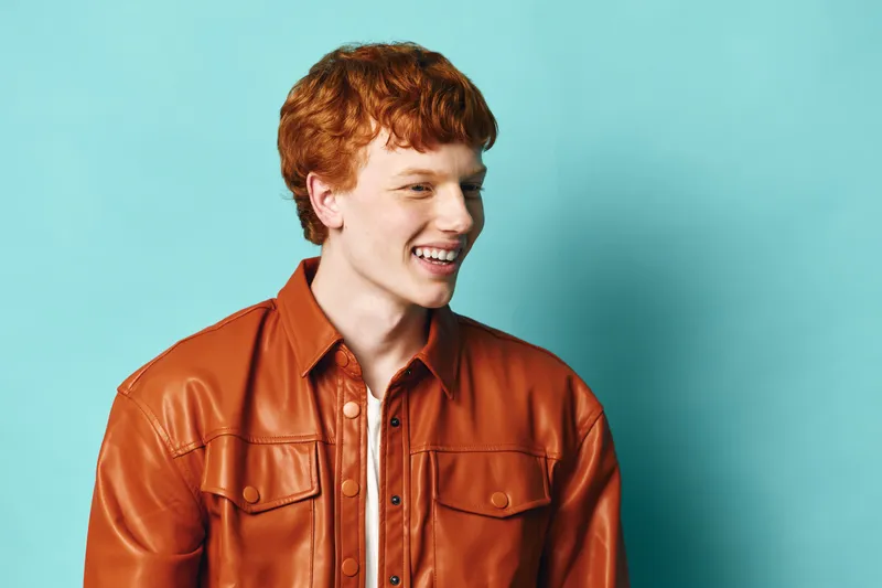 Young man with red curly hair wearing brown leather jacket smiling and looking side on blue background with casual style and happy expression for lifestyle portrait.