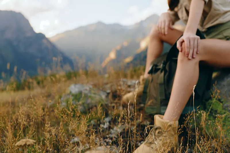 A woman sits on a grassy hillside in outdoor scenery, mountains in the background. Casual hiking attire and boots convey a relaxed, adventurous lifestyle.