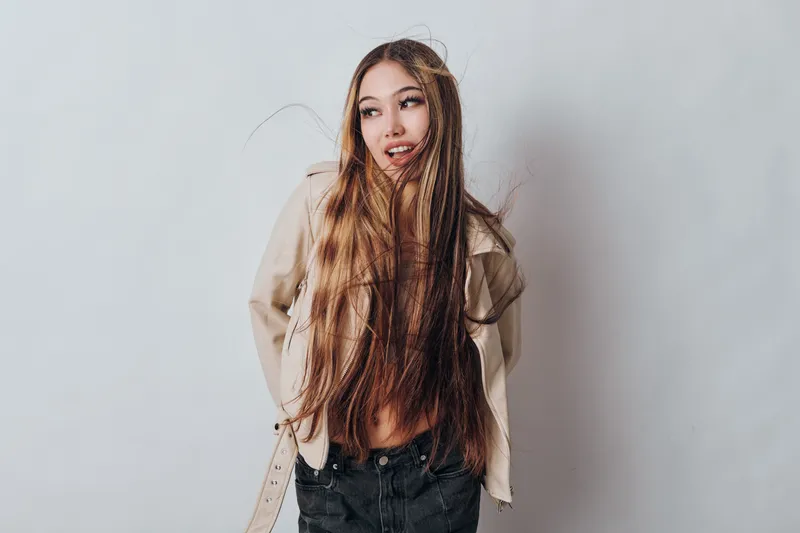 Woman with long hair posing in jacket and jeans standing against light wall. Female model with casual style and relaxed expression looking aside for fashion or lifestyle portrait.