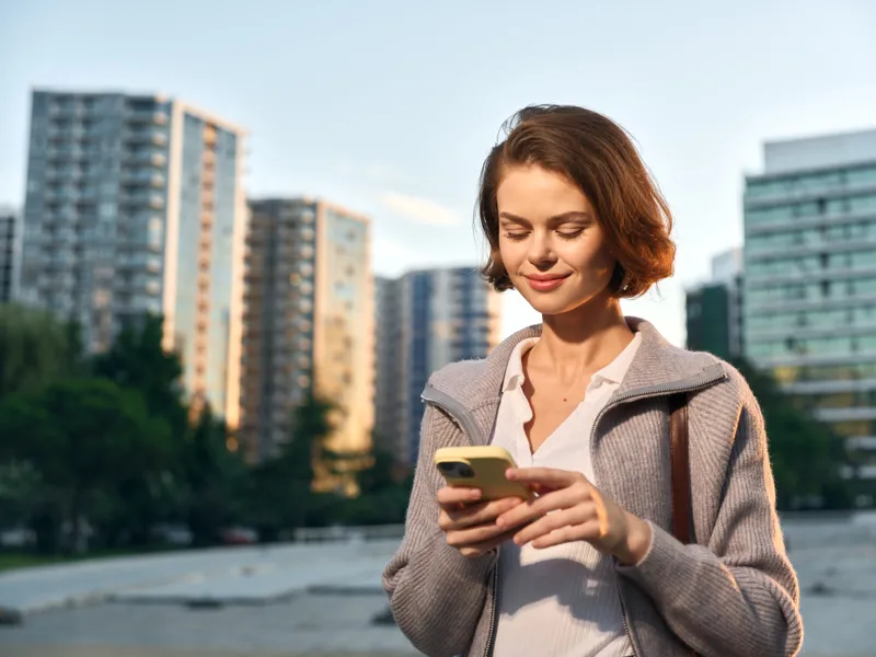 A cheerful young woman in a light coat stands in an urban waterfront park, texting on her smartphone with a focused smile, producing a positive, modern business atmosphere.