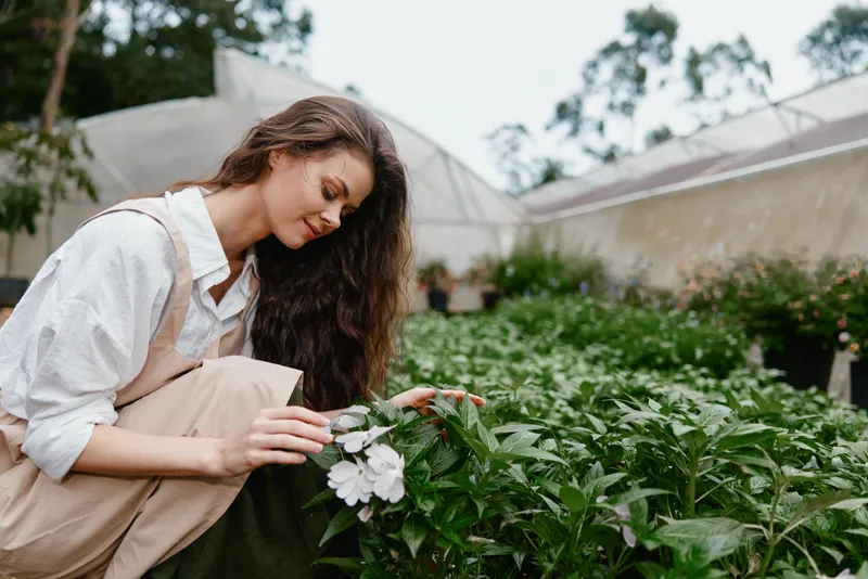 Young woman in beige overalls gently examining flowers in a greenhouse, showcasing a connection with nature and the joy of gardening, ideal for seasonal themes