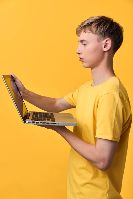 Slim male in a yellow tee holds a laptop in a bright studio, focusing on the screen, showcasing modern technology and remote work in a bold monochrome setting with a clean professional vibe