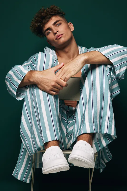 Young man wearing striped pajamas sitting on chair hold coffee mug against dark inclusive modern urban style with relaxed casual feeling and minimalism background in studio