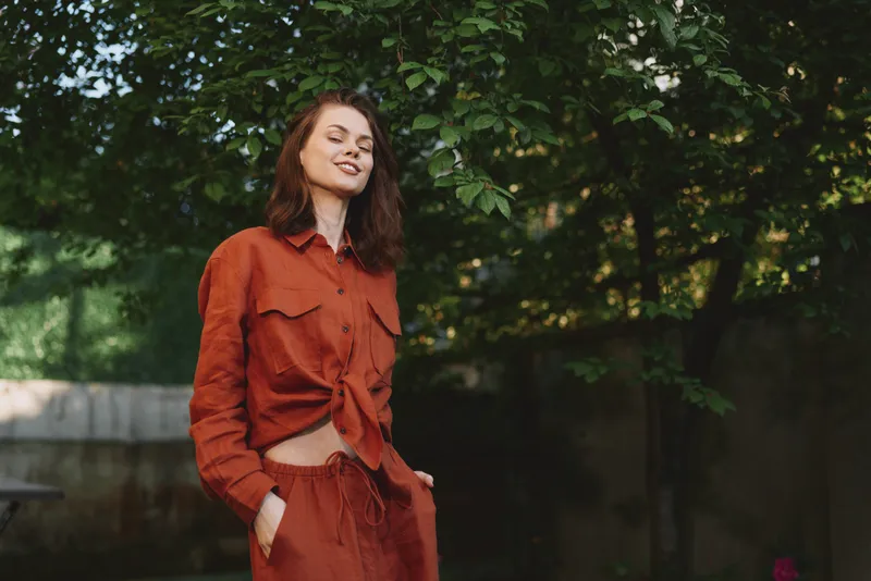 A woman in a rust orange outfit stands outdoors, smiling in a sunlit garden. A casual lifestyle portrait emphasizing fashion, confidence, and relaxed wellness