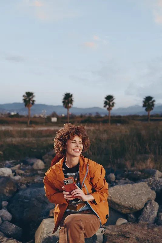Woman lifestyle outdoors, curly hair and orange jacket, sitting on rocks in a field at sunset, smiling while holding a smartphone, soft golden light, casual calm mood, gentle breeze