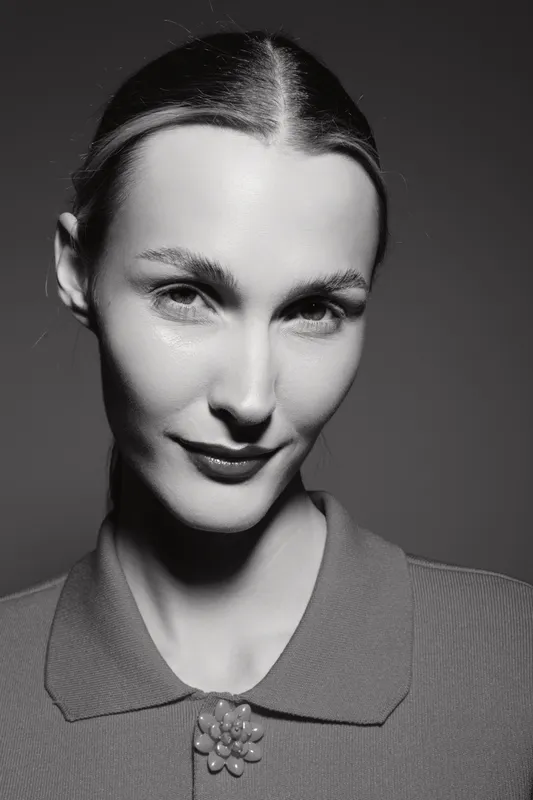 Portrait of a Caucasian woman with pale skin and slicked back hair wearing a collared top with a brooch. Studio lighting highlights facial features in black and white.